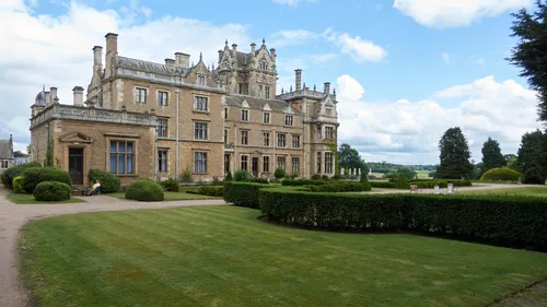 The image shows an impressive view of Thoresby Hall, a grand country house located in Nottinghamshire, England, United Kingdom. Captured on a bright summer afternoon, the photograph highlights the hall's stunning architectural features set against a clear blue sky with scattered clouds. The well-maintained lawn and manicured hedges in the foreground complement the historic building’s detailed stonework and numerous windows, enhancing the stately appearance of this English country house. As a landscape and architectural photograph, the scene conveys the elegance and heritage of Thoresby Hall within its expansive English estate, typical of the historic buildings found in this region of the United Kingdom.